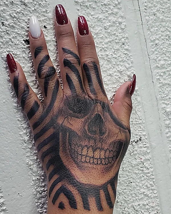 Tatuaje de calavera en negro y gris sobre el dorso de la mano, rodeada de franjas negras en los dedos